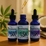 Three blue glass bottles of Balence Hemp CBD Oil labeled 500mg, 1500mg, and 3000mg sit on a smooth wooden surface in warm daylight. The middle bottle’s dropper is raised, releasing a golden drop of oil. Fresh hemp leaves, a halved coconut, and a soft beige background complete the natural, spa-like composition.