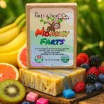 A bar of handmade “Monkey Farts” soap by The Bath & Bodi Co. by Balence sits on a wooden board surrounded by colorful fruit—bananas, kiwi, grapefruit, berries—and small candy cubes. The label shows a playful monkey illustration and rainbow lettering, describing the scent as a fruity blend of banana, grapefruit, kiwi, bubblegum, berries, and vanilla.