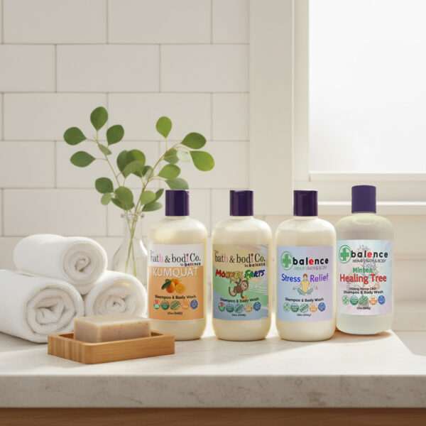 Four Balence shampoo & body wash bottles — Healing Tree, Kumquat, Monkey Farts, and Stress Relief — displayed together on a bathroom counter with towels, eucalyptus sprigs, and a loofah, in bright natural light.