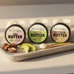 Three Balence Body Butter jars—Shea Butter, Avocado Butter, and Grass-Fed Beef Tallow Butter—arranged on a rustic wooden tray with soft linen, natural light, and accents of shea nuts, avocado slices, and fresh green grass for a pure spa-like display.
