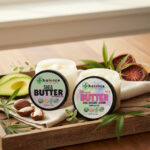 Two Balence Hemp Shea Butter jars—classic Shea Butter with hemp CBD and Ultimate Butter (Shea, Avocado, Kokum, 250 mg hemp CBD)—side by side on a rustic wooden tray with shea nuts, fresh avocado slices, kokum fruit, and hemp leaves, softly lit by morning sunlight for a clean, spa-like vibe.