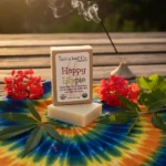 Balence Happy Hippie handmade soap bar on rustic wood with tie-dye cloth, hemp leaves, geranium flowers, patchouli sprigs, and incense smoke in warm sunlight.