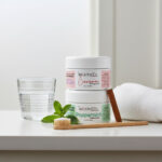 Two Balence Bentonite Clay Tooth Powder jars (Cinnamon and Peppermint) on a bathroom counter with a bamboo toothbrush coated in tooth powder resting on a dish. Fresh mint, a cinnamon stick, glass of water, and towel complete the natural, refreshing scene.