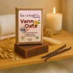 Balence Vann Oats non-CBD bar soap rests on a wooden bath tray surrounded by vanilla beans, oat flakes, and a softly glowing candle. A rolled towel and delicate oat sprigs create a warm, serene spa-like atmosphere with natural, earthy tones.